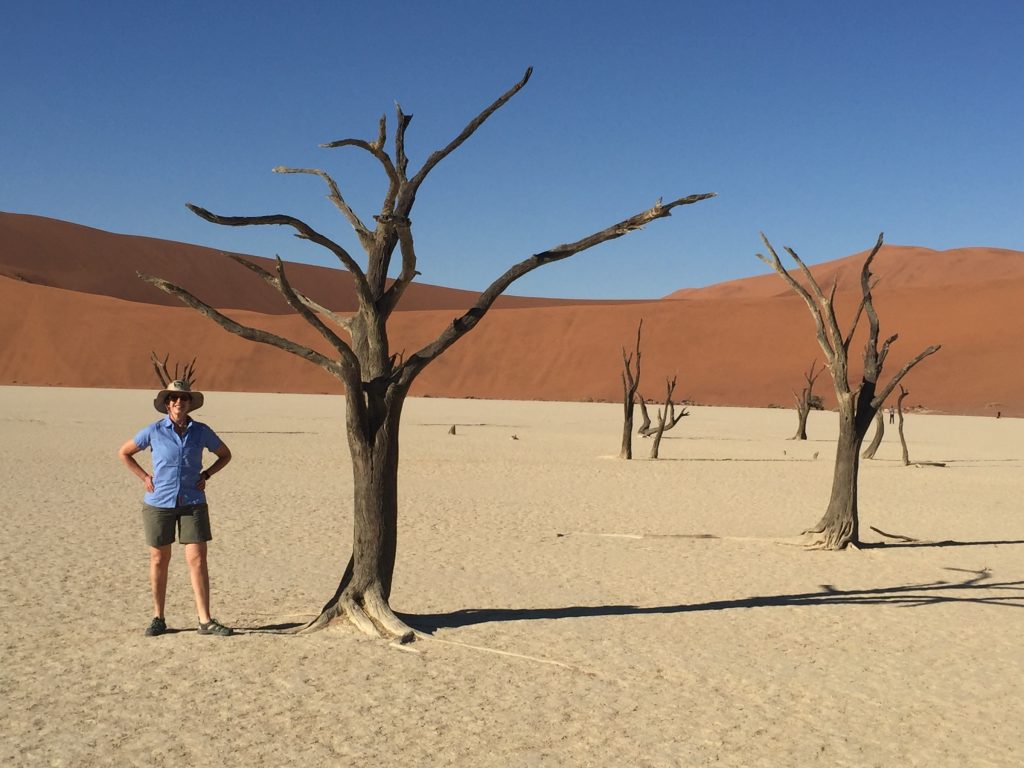 Photo: Dr. Beth Karlan wearing safari hat, shorts and sunglasses, standing next to Camel Thorn Trees